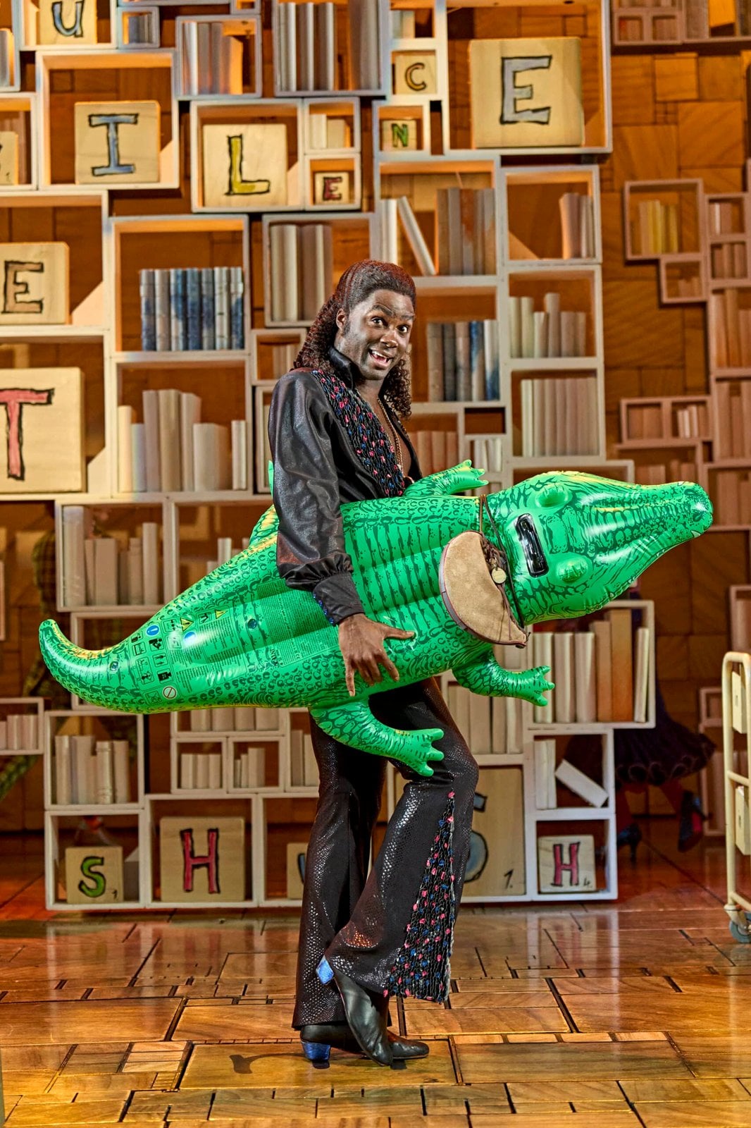 Performer holding a green crocodile prop against a bookcase set